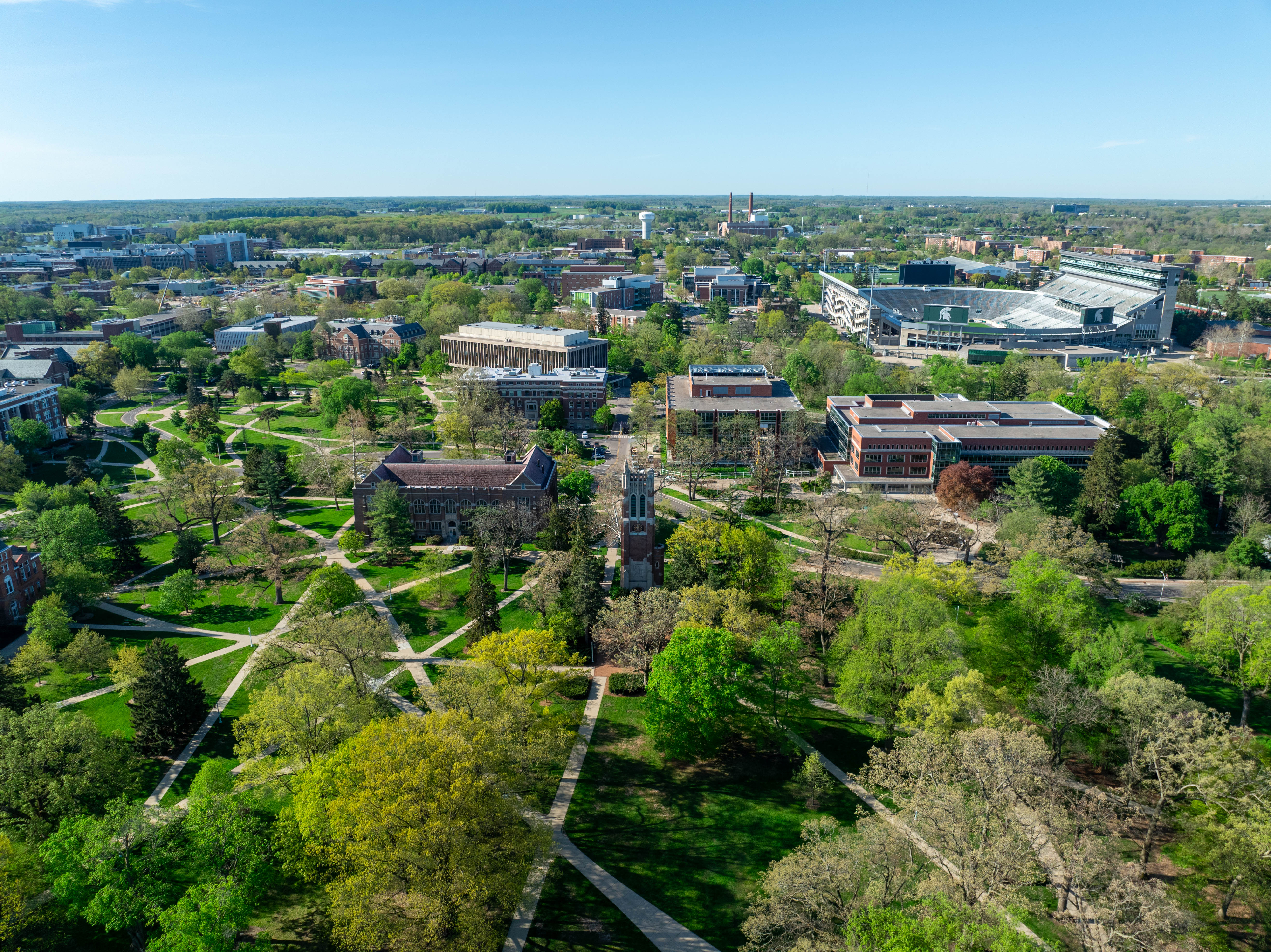 Aerial view of Michigan State University’s campus on a bright spring day, showing green trees, walking paths across the open lawns, academic buildings, Beaumont Tower near the center, and Spartan Stadium visible in the distance.