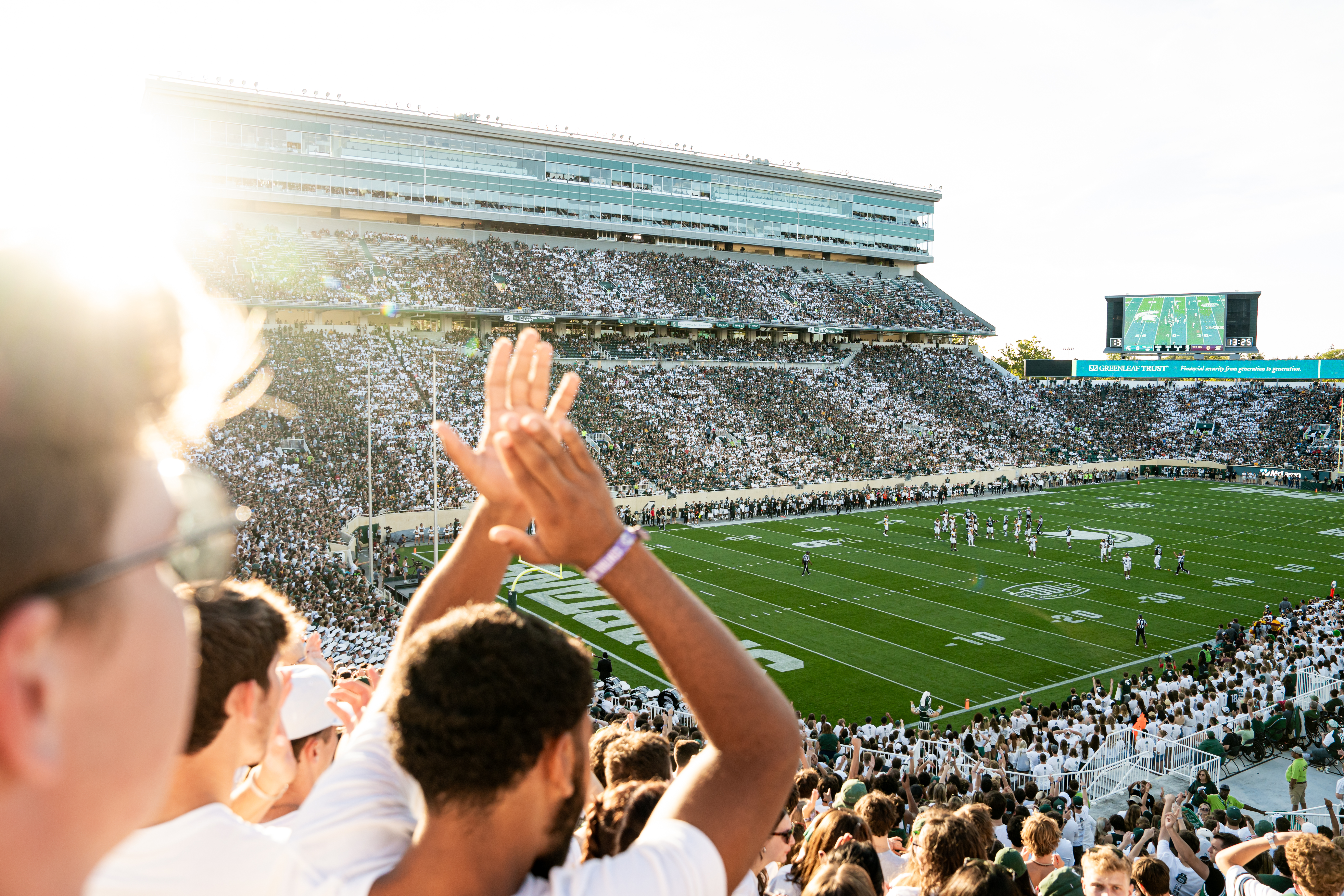 Wide view of Michigan State University’s Spartan Stadium during a football game, with fans in the foreground cheering and clapping while players line up on the field under bright afternoon sunlight.