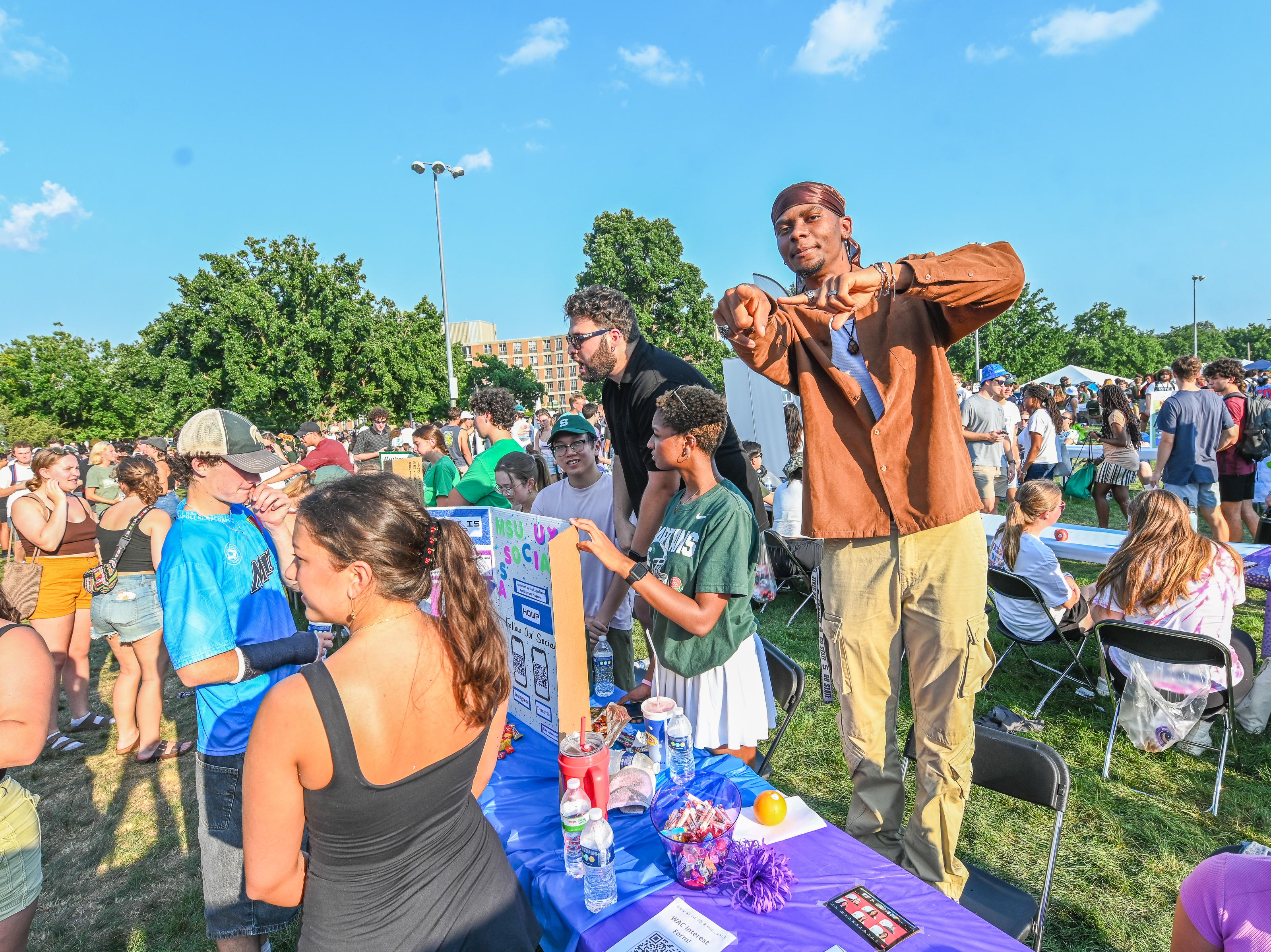 Students gather outdoors at an MSU event on a sunny day, exploring tables covered with displays, flyers, and promotional items. People walk through the crowded area, talk with student organizations, and participate in activities under a bright blue sky.