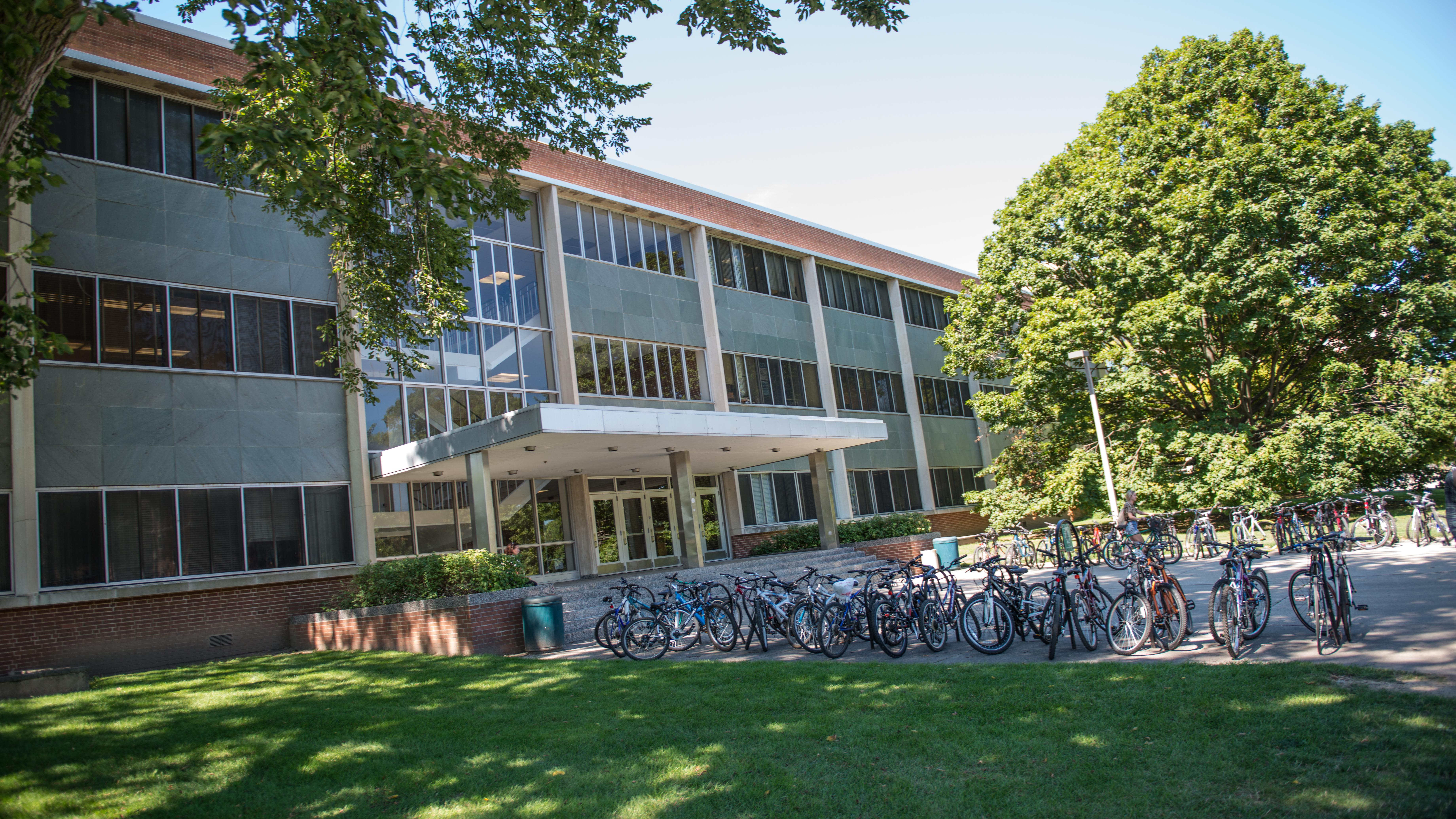 A large academic building with multiple floors and tall windows sits behind a wide bike rack filled with many bicycles. The building has a covered entrance supported by pillars and is surrounded by green lawns, mature trees, and landscaped shrubs. The scene is bright and sunny with clear blue skies.