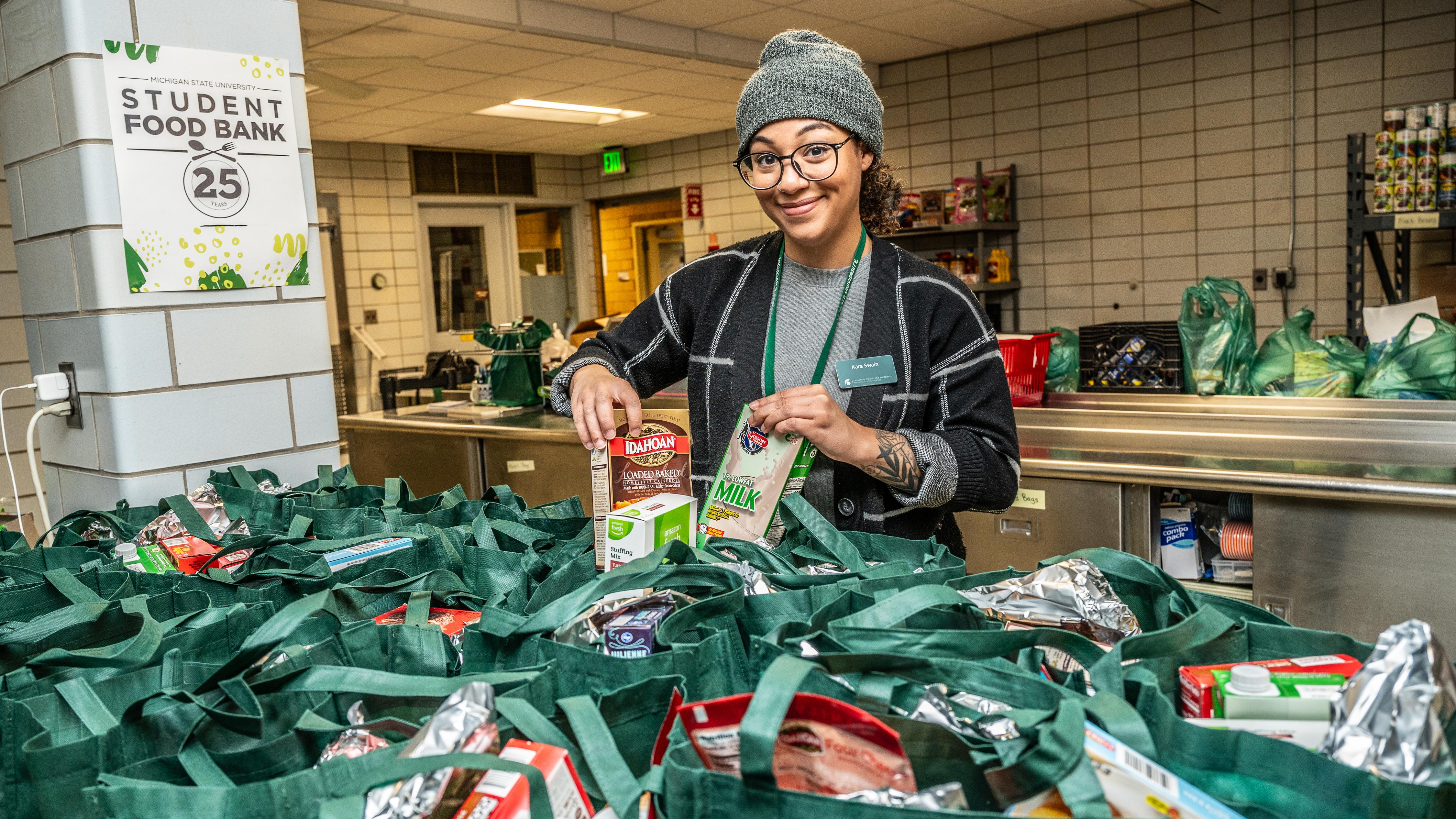 A person stands behind a counter assembling green reusable grocery bags filled with pantry items such as boxed oatmeal, cartons of milk, and canned goods. The setting appears to be a campus food bank, with shelves of supplies in the background and a sign on the wall reading “Student Food Bank.” The individual is sorting items into the bags, which are lined up and filled with various packaged foods