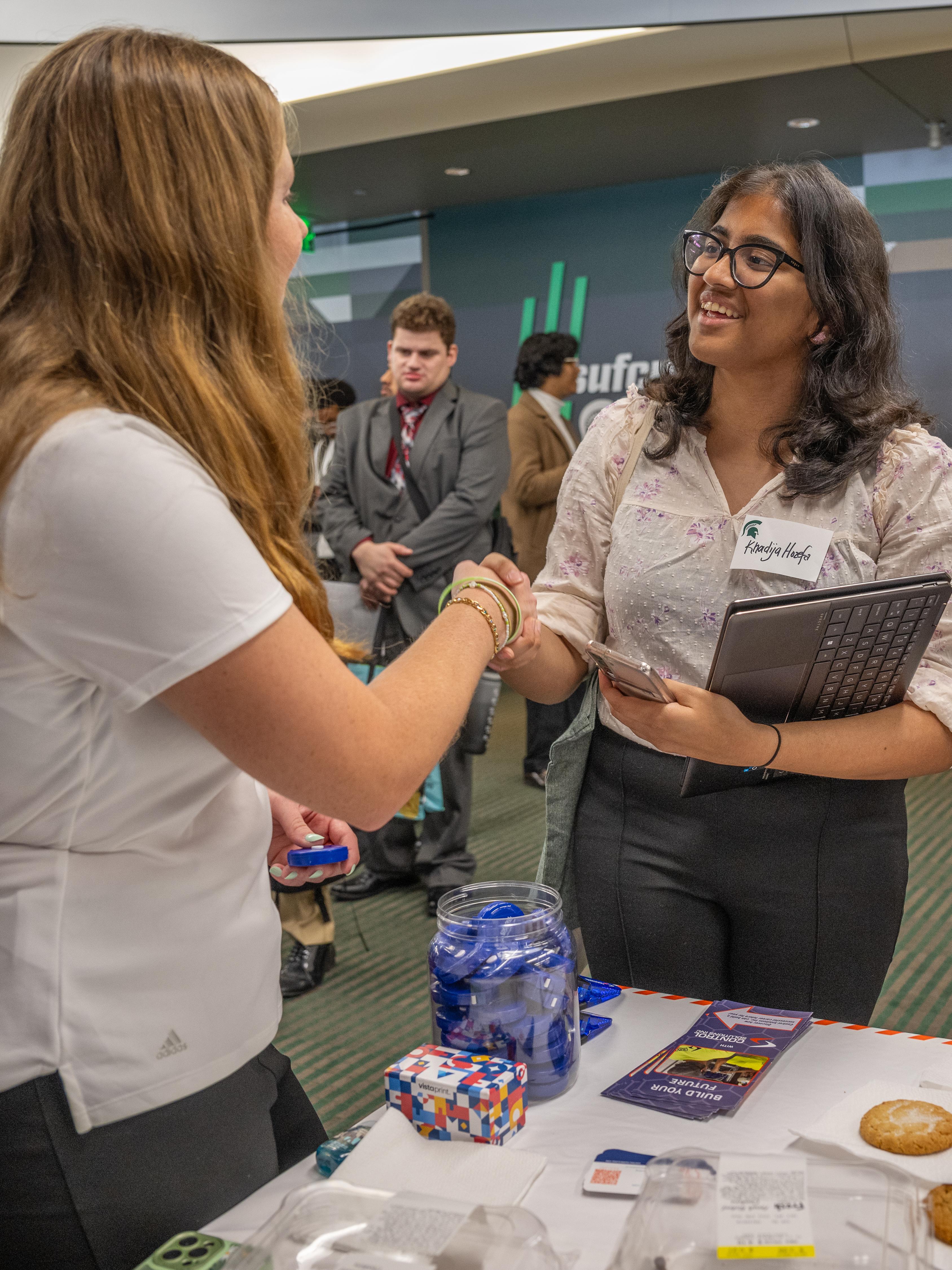Two individuals shake hands at a professional event while standing at a table displaying promotional materials, small giveaway items, and a jar filled with branded items. One person holds a tablet and a phone, and several other attendees converse in the background within a modern indoor venue