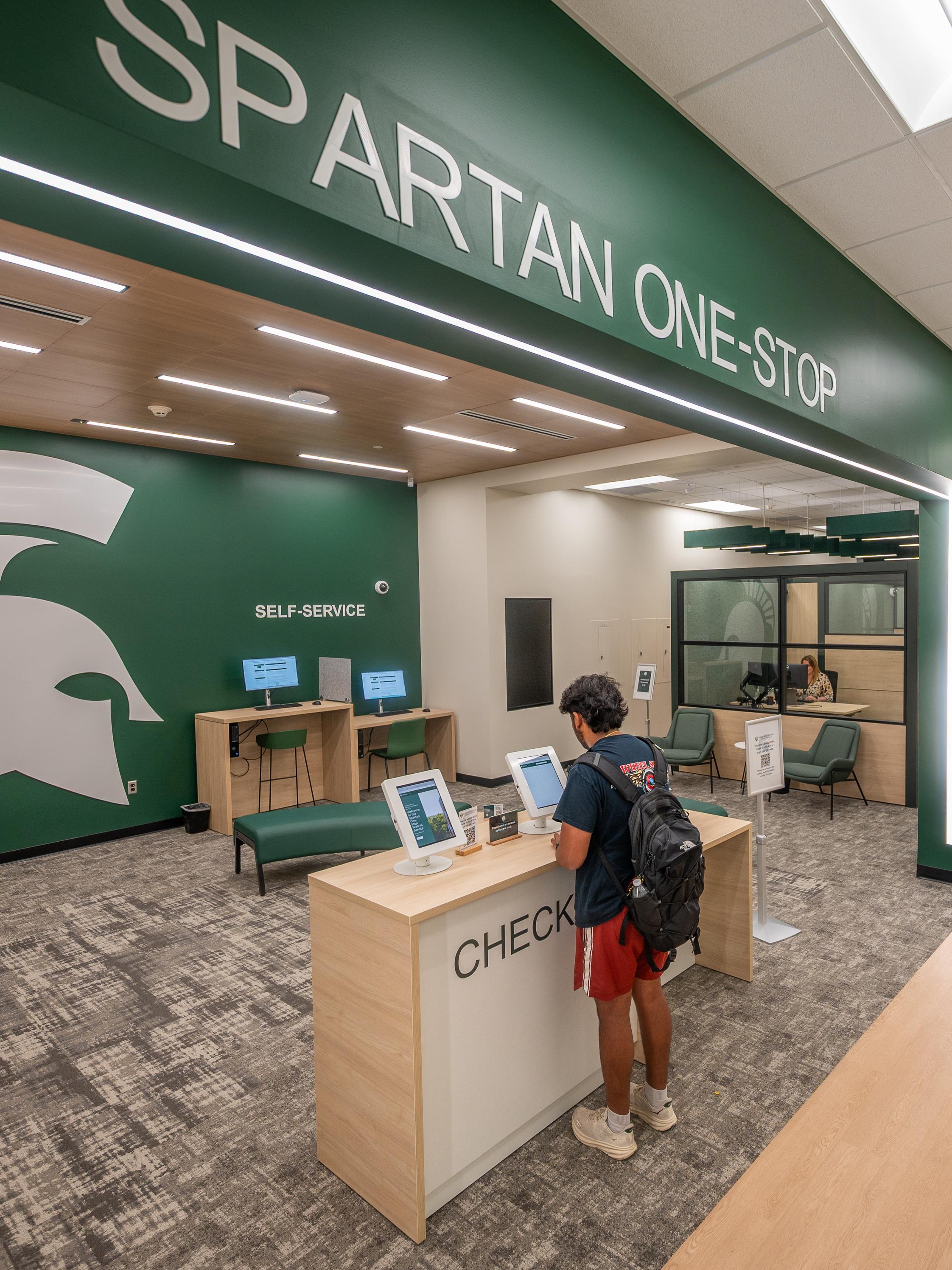 A student stands at a check-in desk inside the Spartan One‑Stop center, a modern space with green walls and the Spartan helmet logo. The area includes self‑service computer stations on the left and private service offices on the right. Bright overhead lighting and wood‑tone flooring create an open, welcoming environment, with a hallway extending along the right side of the image.